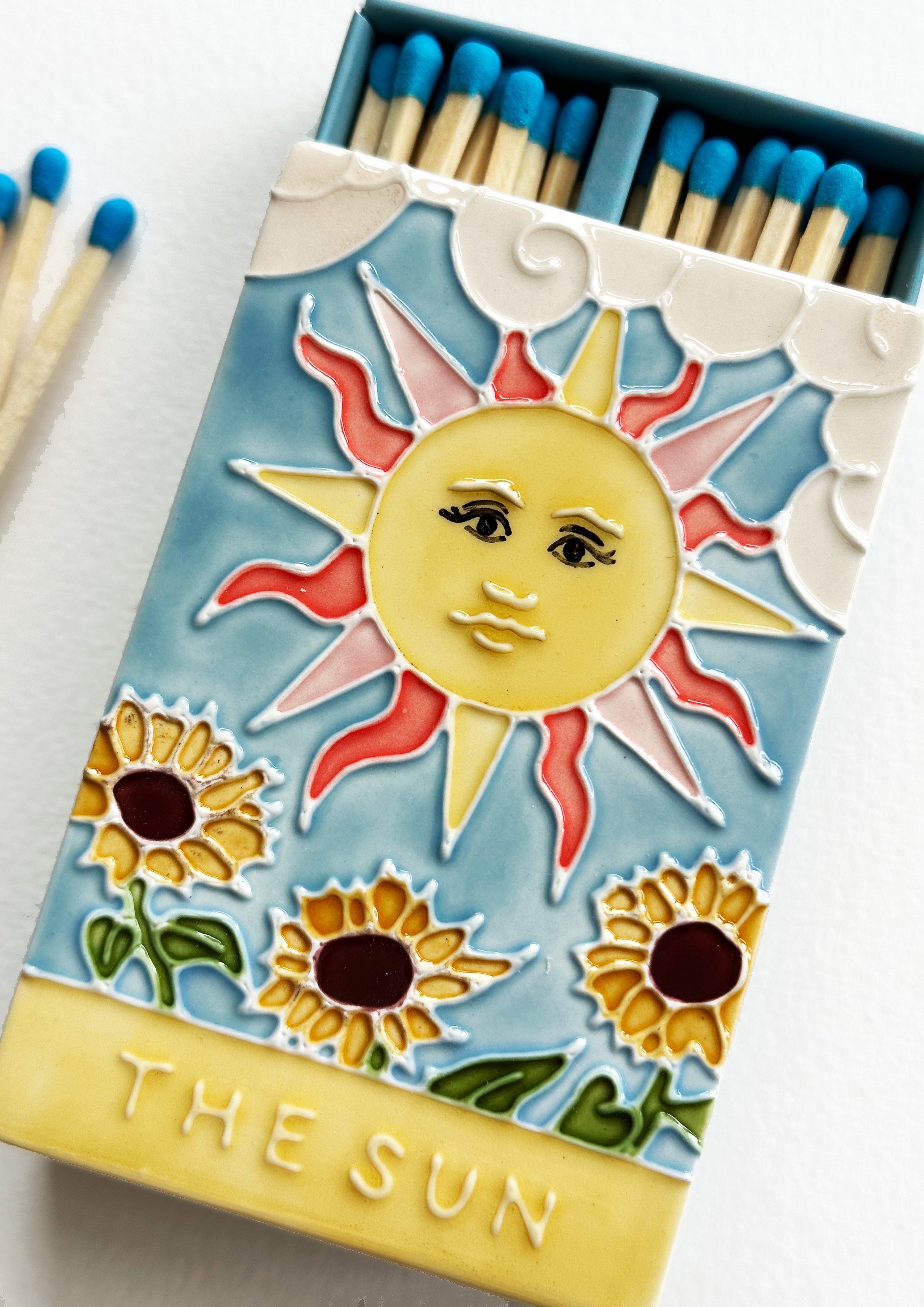 Luxury ceramic matchbox featuring The Sun Tarot card - a full shining sun sits above a sunflower field. The ceramic artwork sits atop a luxury card case with striker strip. Each box contains 40 aqua coloured extra long safety matches. Made exclusively in England.