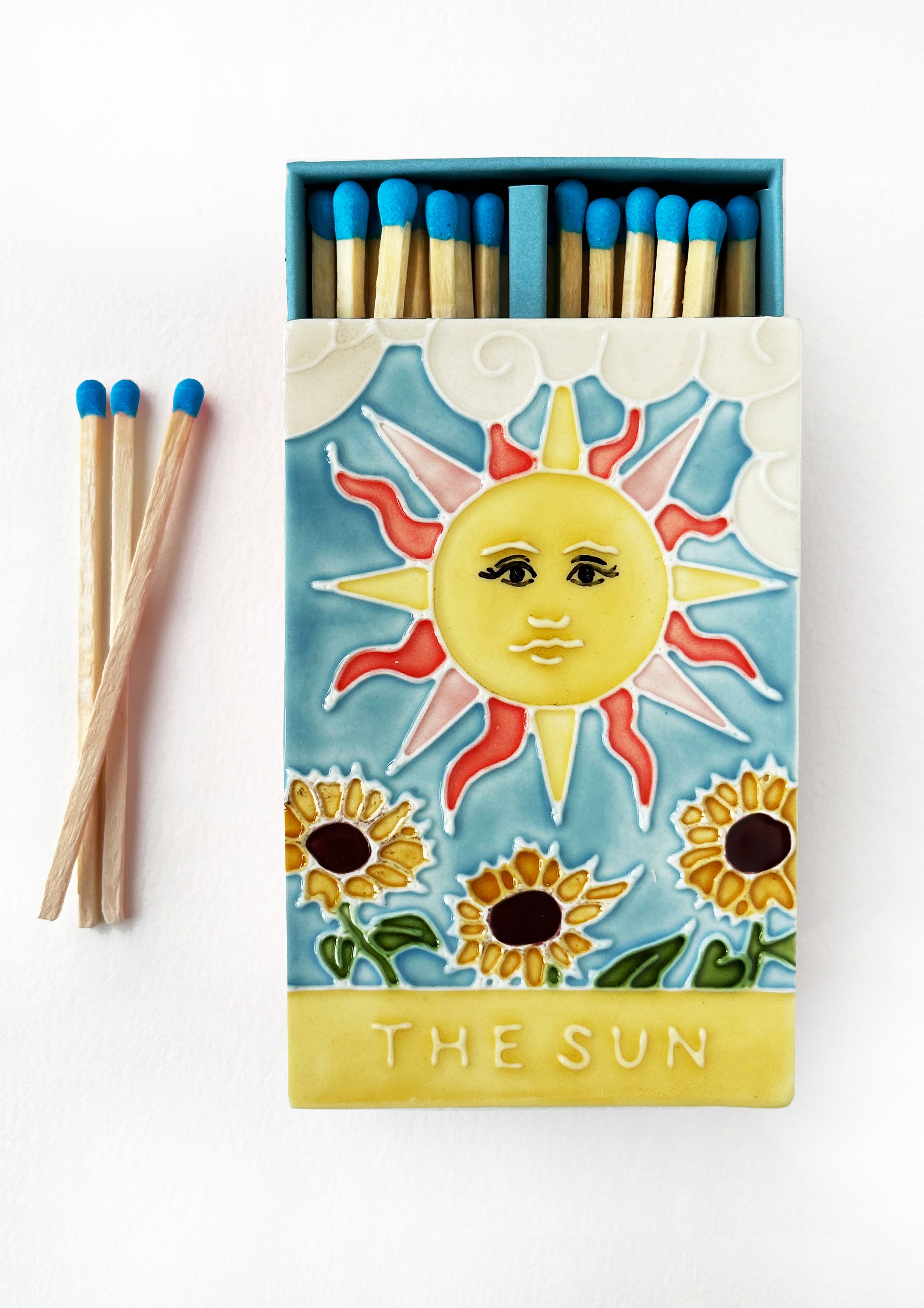 Luxury ceramic matchbox featuring The Sun Tarot card - a full shining sun sits above a sunflower field. The ceramic artwork sits atop a luxury card case with striker strip. Each box contains 40 aqua coloured extra long safety matches. Made exclusively in England.