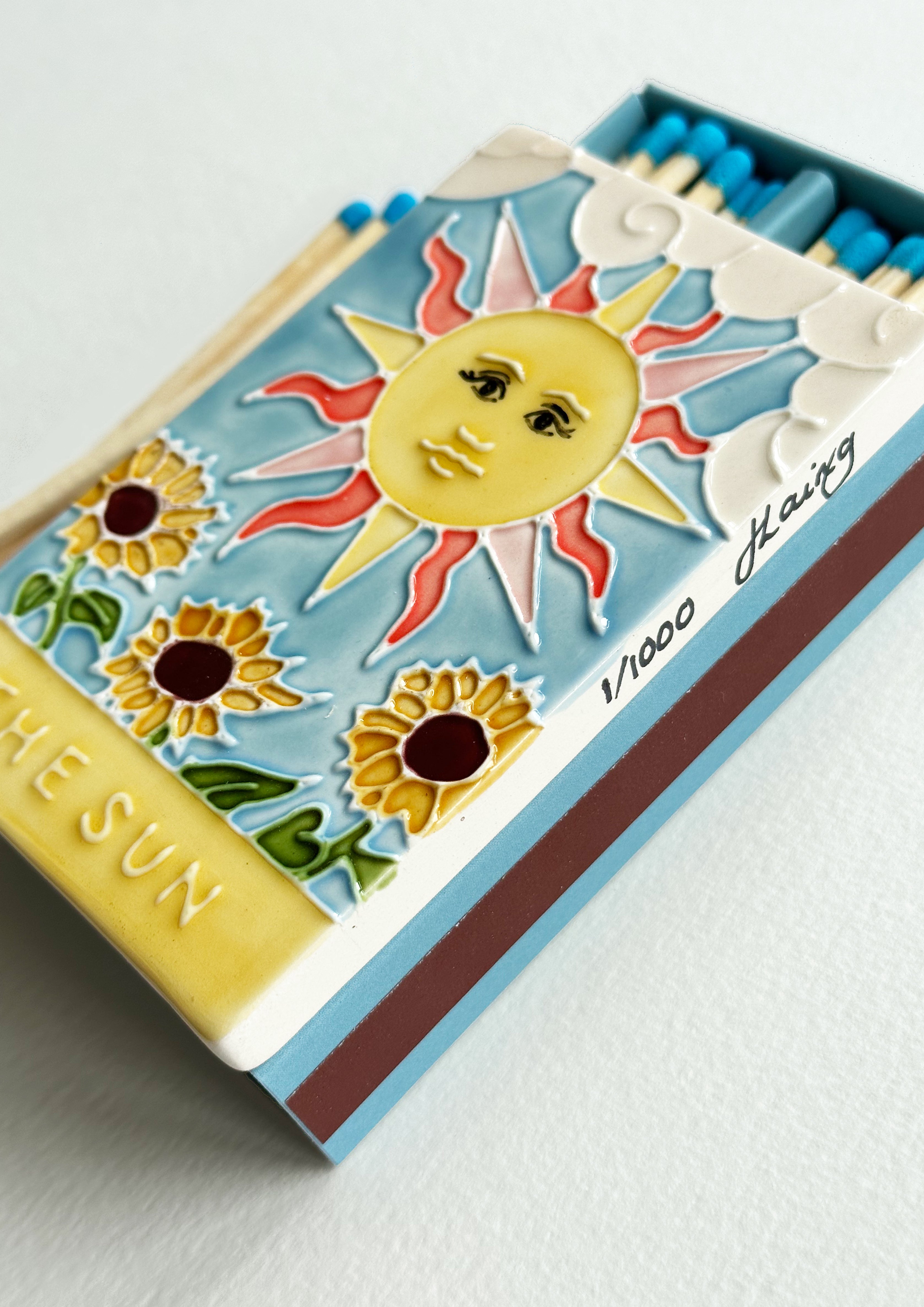 Luxury ceramic matchbox featuring The Sun Tarot card - a full shining sun sits above a sunflower field. The ceramic artwork sits atop a luxury card case with striker strip. Each box contains 40 aqua coloured extra long safety matches. Made exclusively in England.