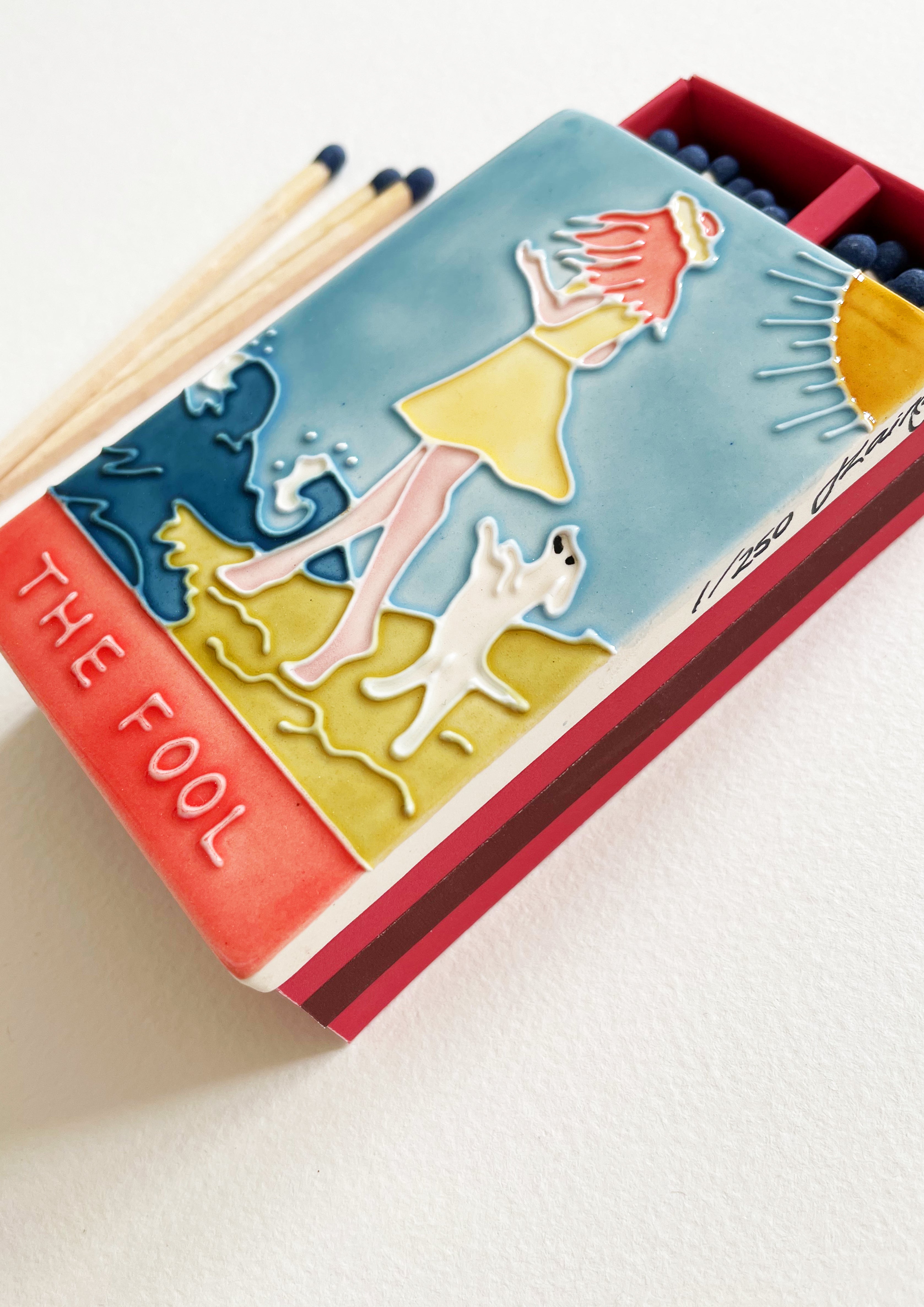 Luxury ceramic 'Moi et Toi' matchbox featuring the Fool tarot card. The ceramic artwork sits atop a luxury card case with striker strip. Each box contains 40 navy coloured extra long safety matches. Made exclusively in England.