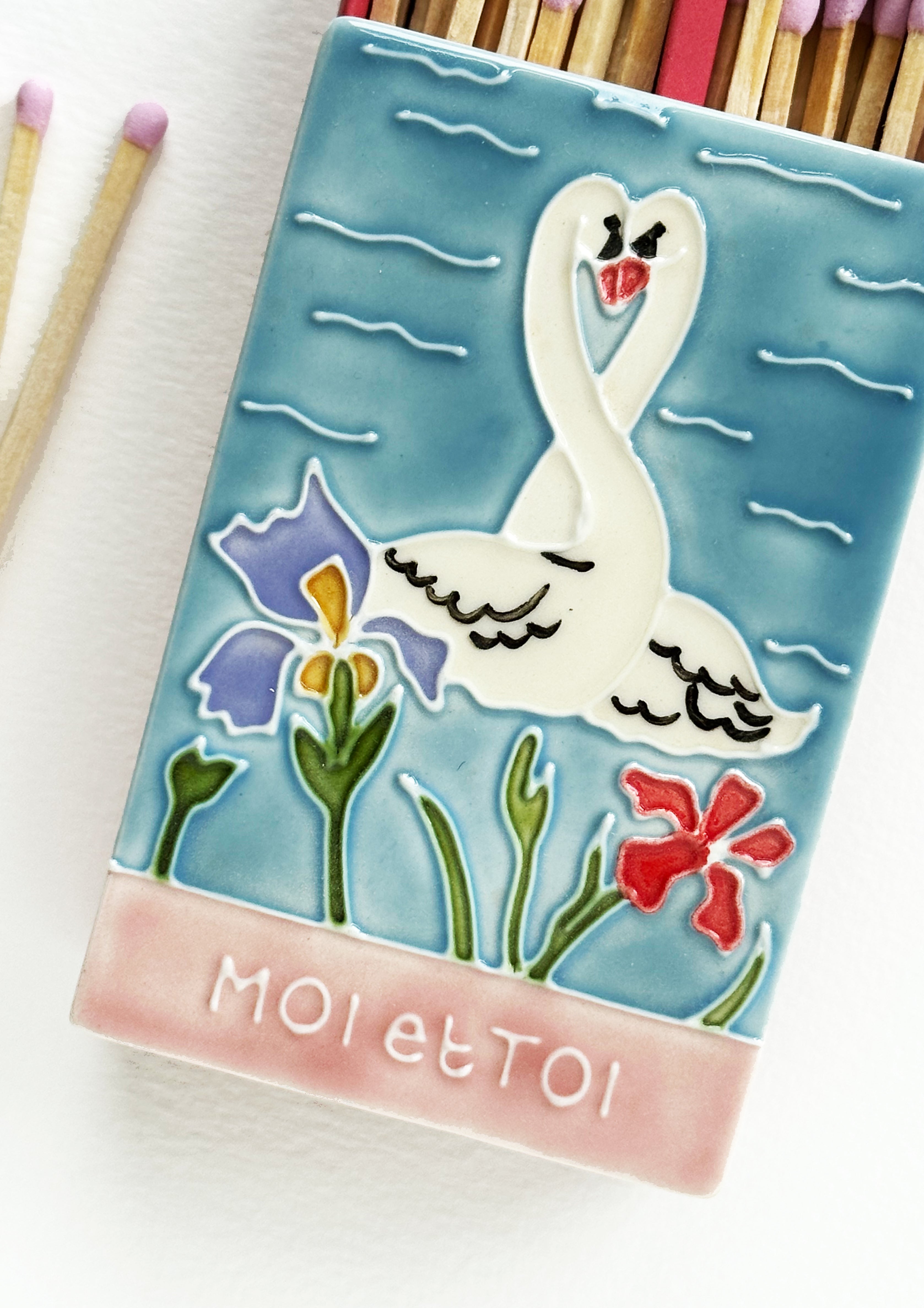 Luxury ceramic 'Moi et Toi' matchbox featuring kissing swans and iris flowers. The ceramic artwork sits atop a luxury card case with striker strip. Each box contains 40 coloured extra long safety matches. Made exclusively in England.